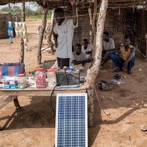 A group of refugees utilizing renewable energy under a hut equipped with a solar panel.