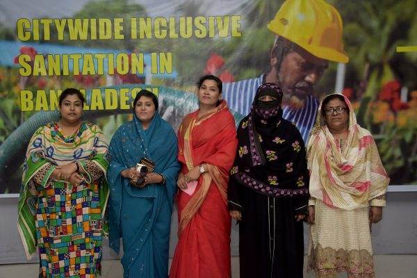 A group of women standing in front of a banner with the words citywide inclusive sanitation in Bangladesh, enjoying a warm and thoughtful breakfast with the WASH ladies!