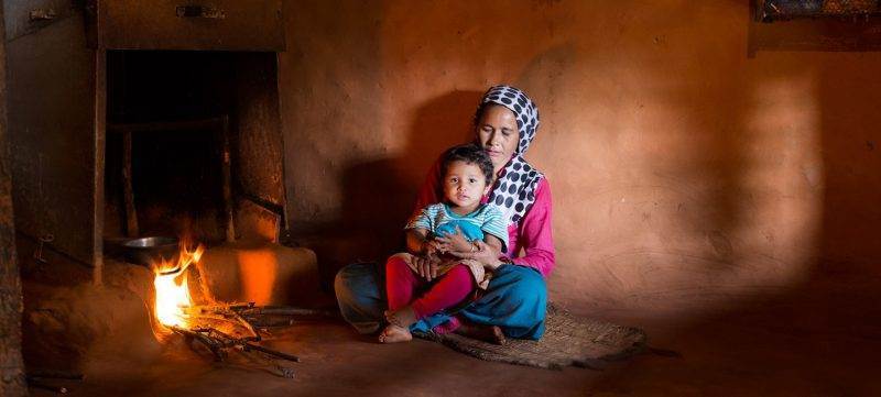 A woman and child sitting in front of a fire in a hut, unaware of the killer in the kitchen.