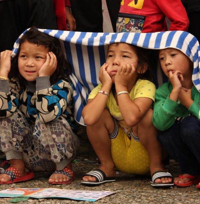 A group of children participating in the Nepal Reconstruction Campaign sit under a blanket.