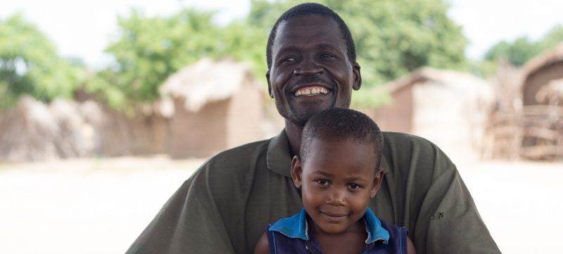 A man **updating his details** while holding a child in front of a hut.