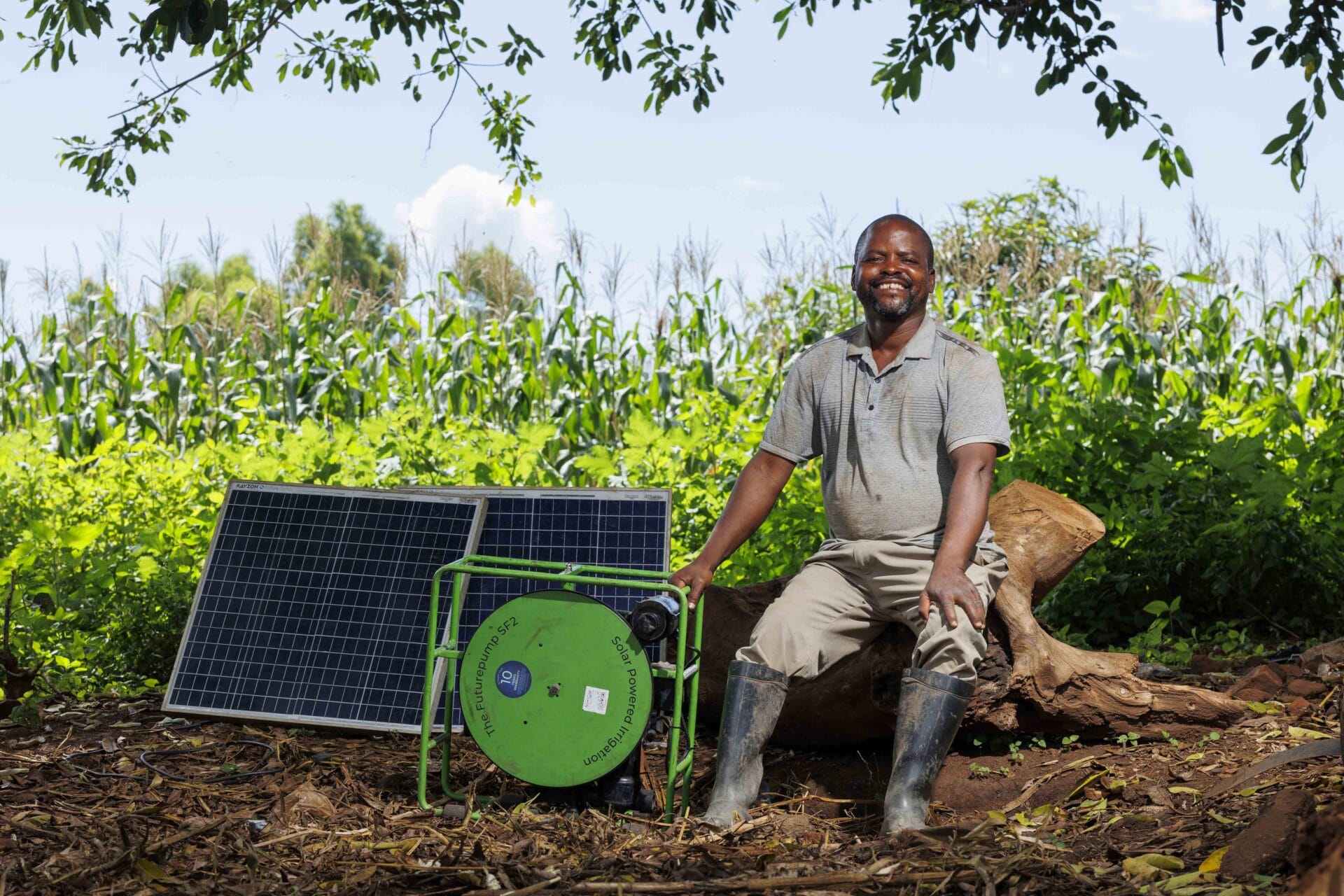 Smiling man in wellies sits by a solar panel and green device in a lush, green farm setting.