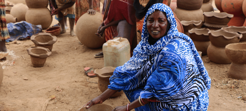 A woman in a blue shirt is making clay pots to clear the air in Darfur.