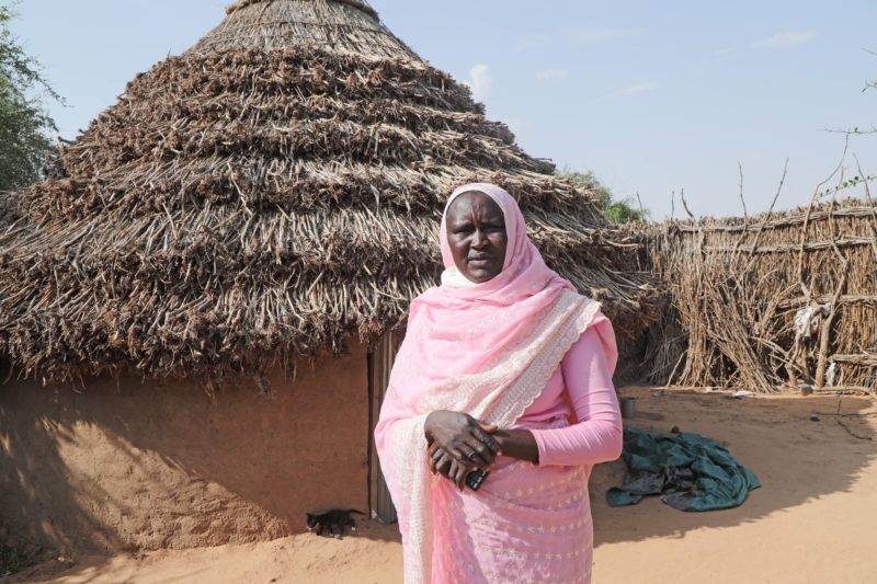 A woman in a pink sari standing in front of a thatched hut symbolizes hardship and hope in North Darfur.