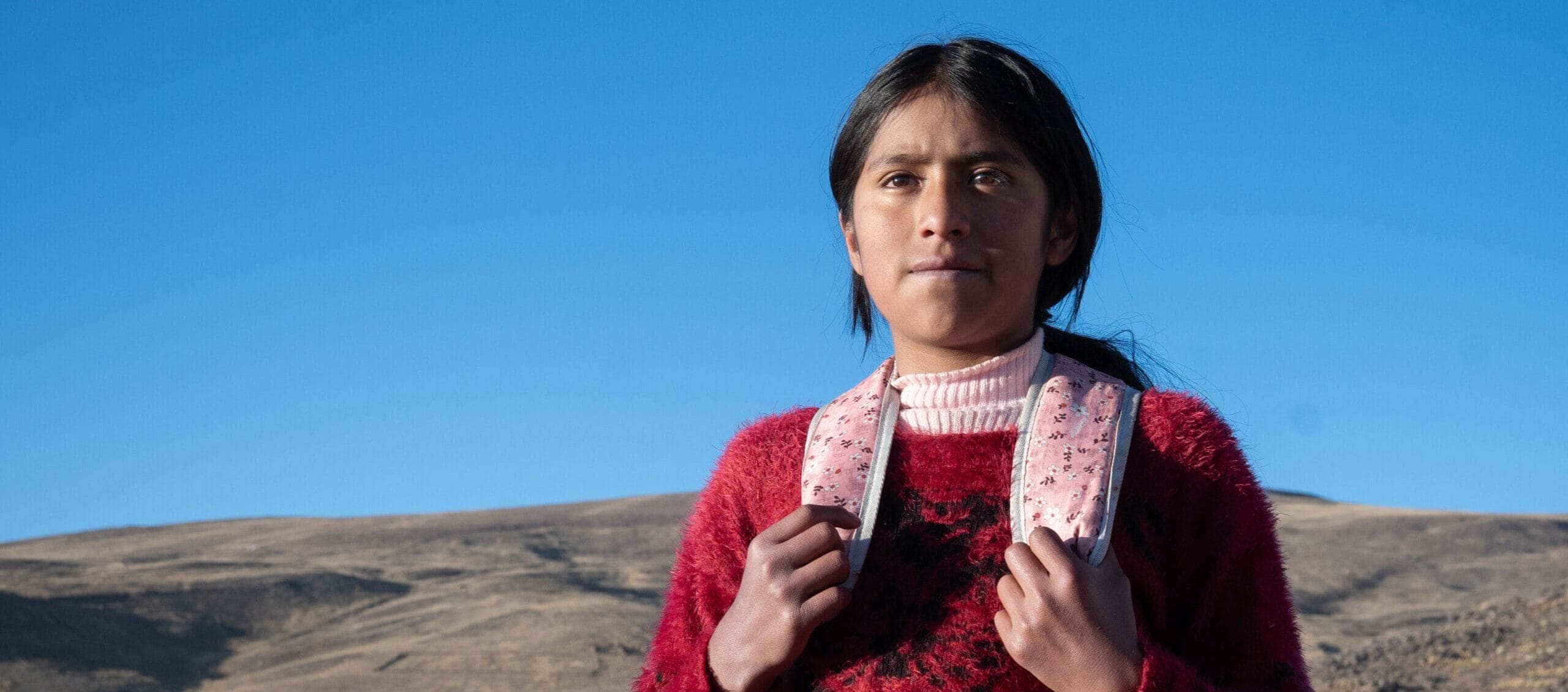 Girl in a red jumper stands outside, holding rucksack straps, with hills and blue sky in the background.