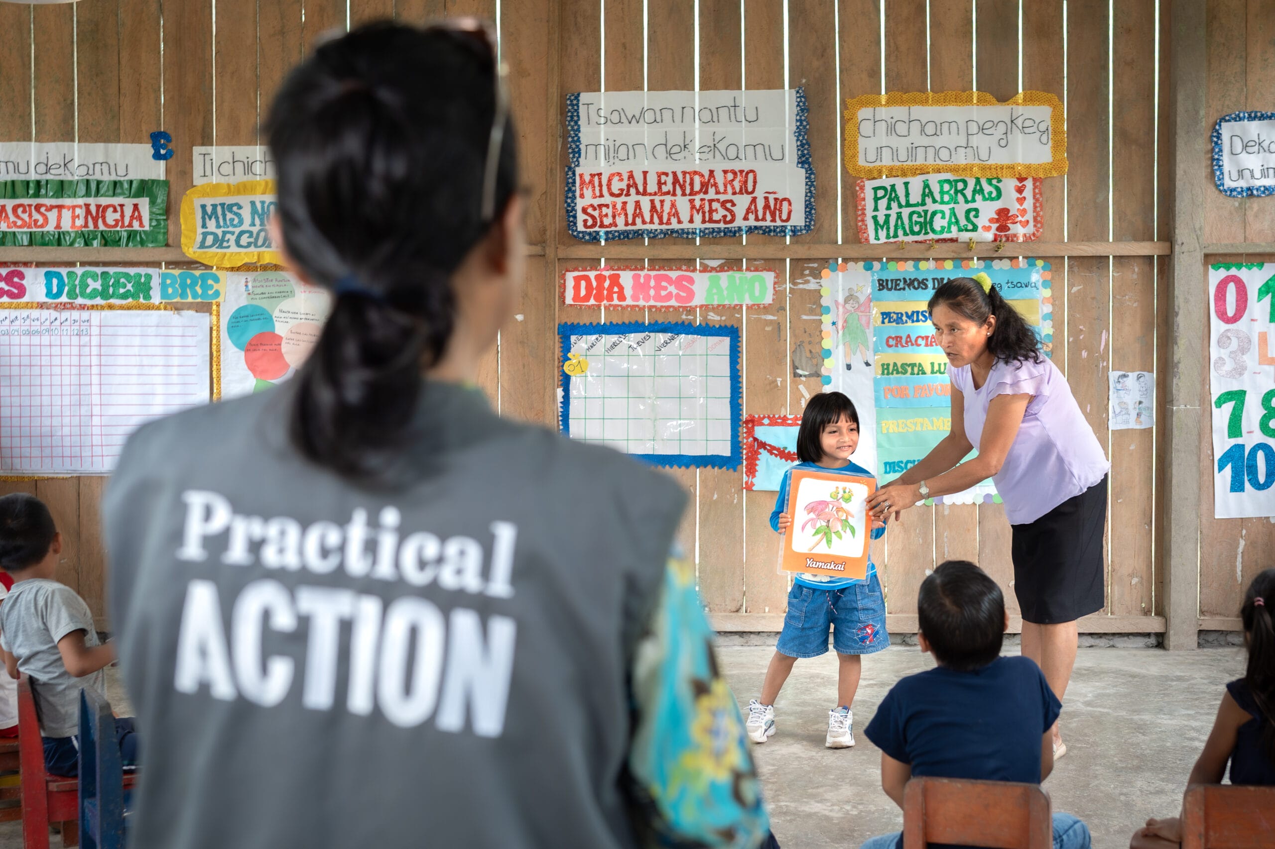 A teacher helps a child display a picture in a classroom, as pupils watch and a person in a “Practical Action” vest observes.