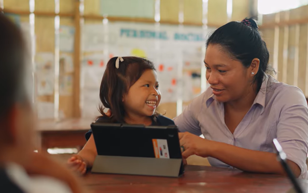 A woman and a young girl smile whilst using a tablet together at a wooden table in a classroom.