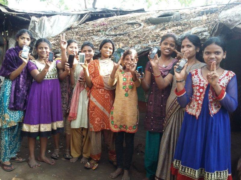 A group of women using cell phones to facilitate gender transformative change in front of a hut.