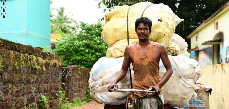 A man cycling with a substantial load of waste on his back.