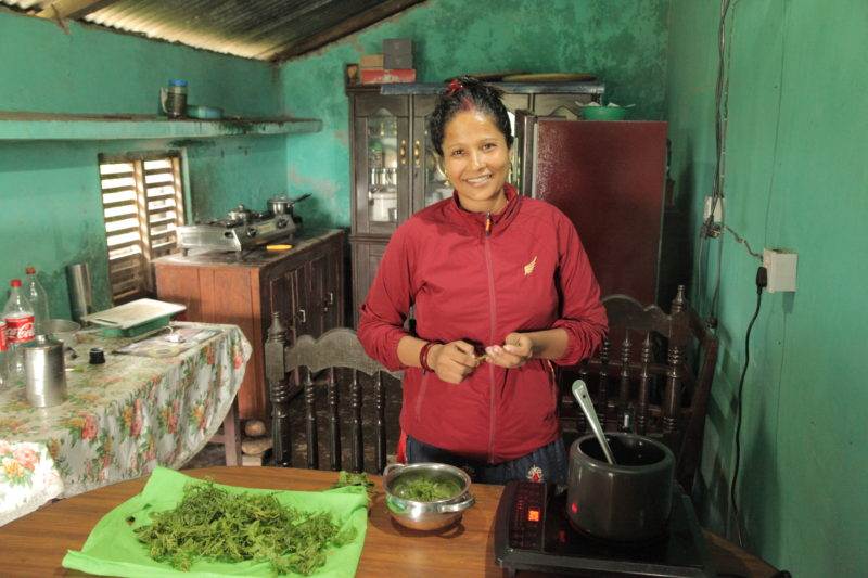 A woman in a red jacket standing in front of a kitchen, involved in a decade-long project funded for clean cooking in Nepal.