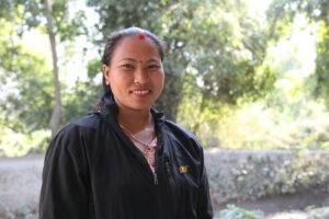 A Nepalise lady called Bideshini Tharu wearing a black jacket over a colourful blouse, a necklace and smiling - stood in a rural area surrounded by trees and shrubs.