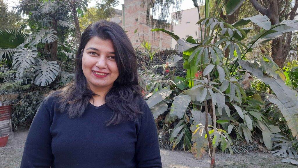 Richa - A Nepalise lady with long black hair, black blouse, smiling whilst stood in a courtyard surrounded by tropical trees and plants