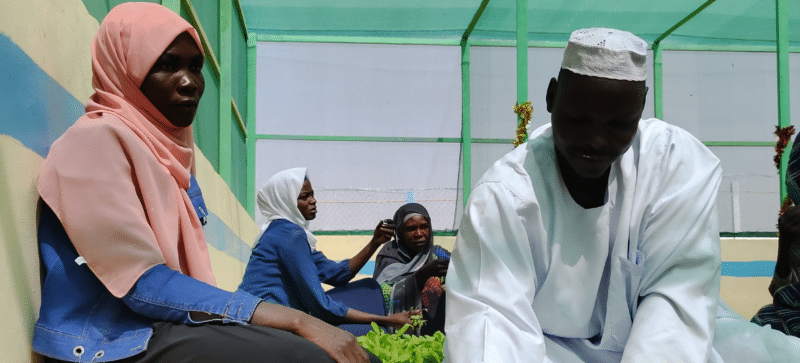 An update from our Sudan director, Muna Eltahir - people sitting on the ground in front of green building.