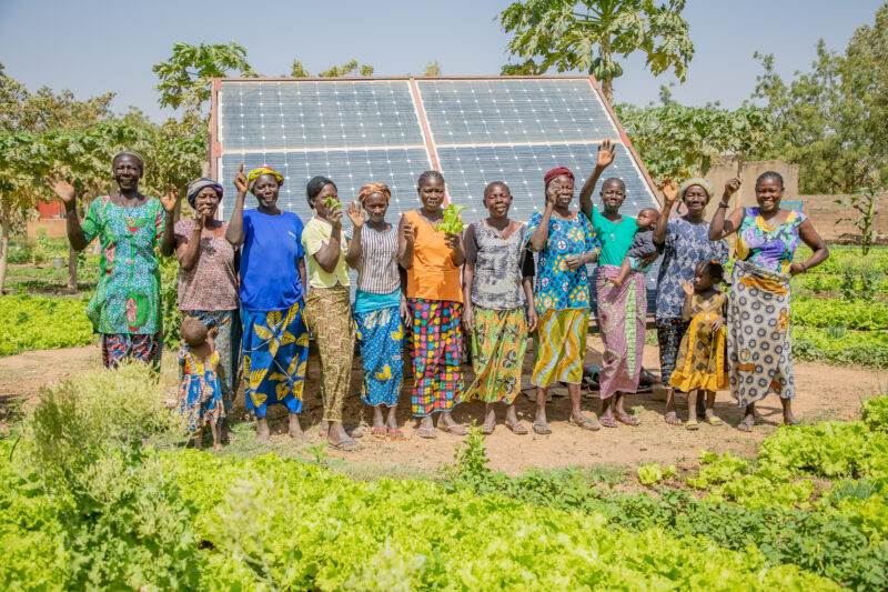 A group of women, seeding transformation in Burkina Faso, standing in front of a solar panel.