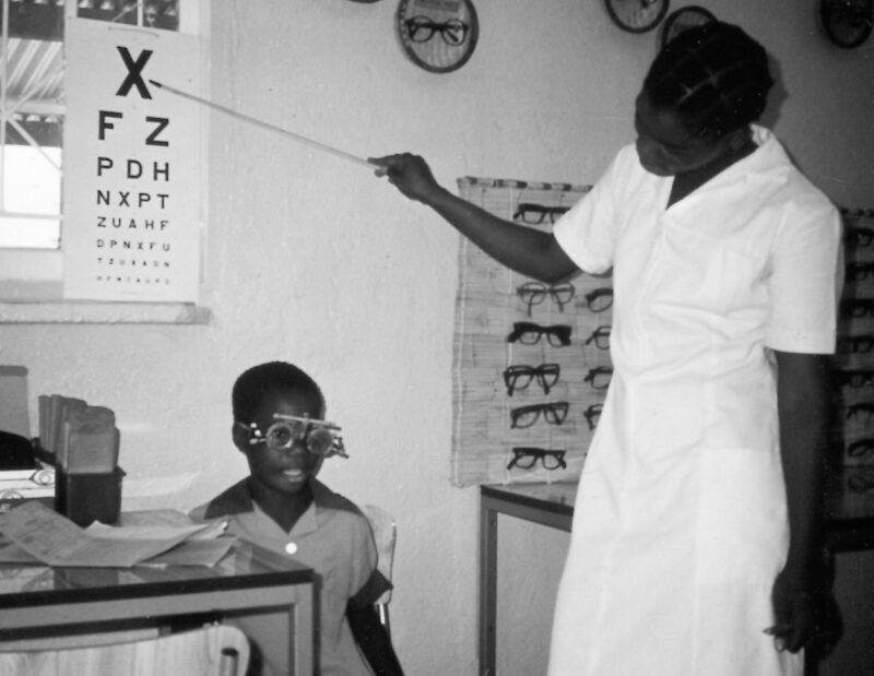 An old black and white photo capturing a woman teaching a child how to read an eye chart, showcasing the beauty of small moments passed by half a decade on.