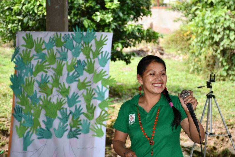 A woman in a green shirt is holding a microphone in front of a tree at the Latin America and Caribbean Climate Week.