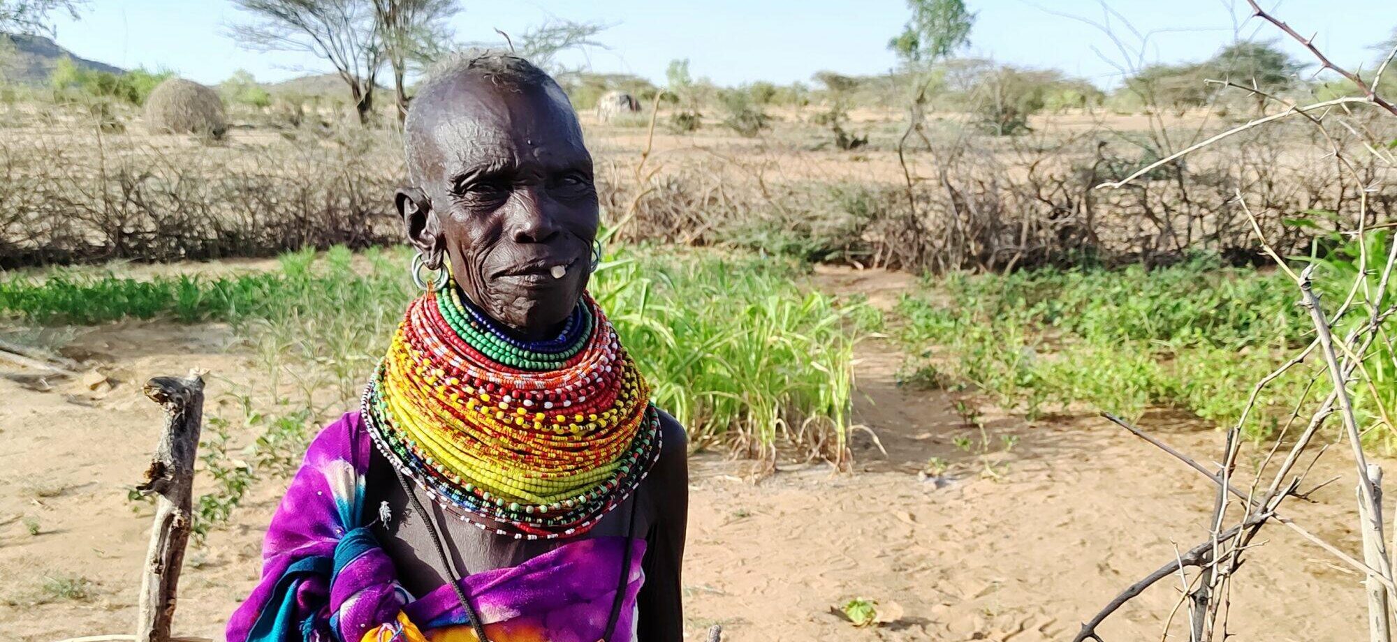 In front of a field that has been sparsely cropped due to the scarcity of water, stands a woman adorned in a vibrant and eye-catching dress.