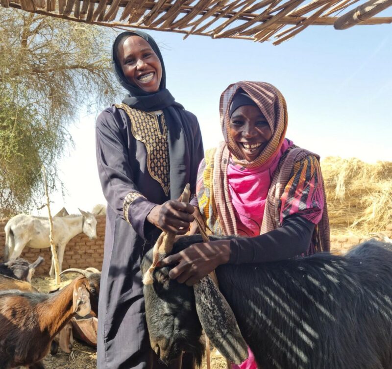 Two women standing next to a goat, symbolizing growing prosperity in North Sudan.