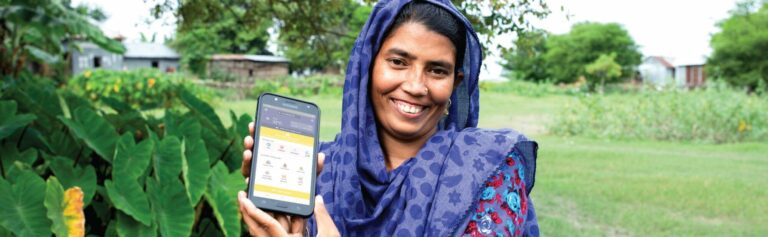 A woman holding up a cell phone in a field, demonstrating Practical Action in Bangladesh.