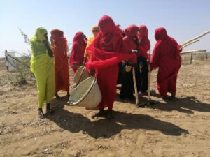 A group of women from the Kassala Women's Development Association Network, wearing red sarongs, stand together in a dirt field.
