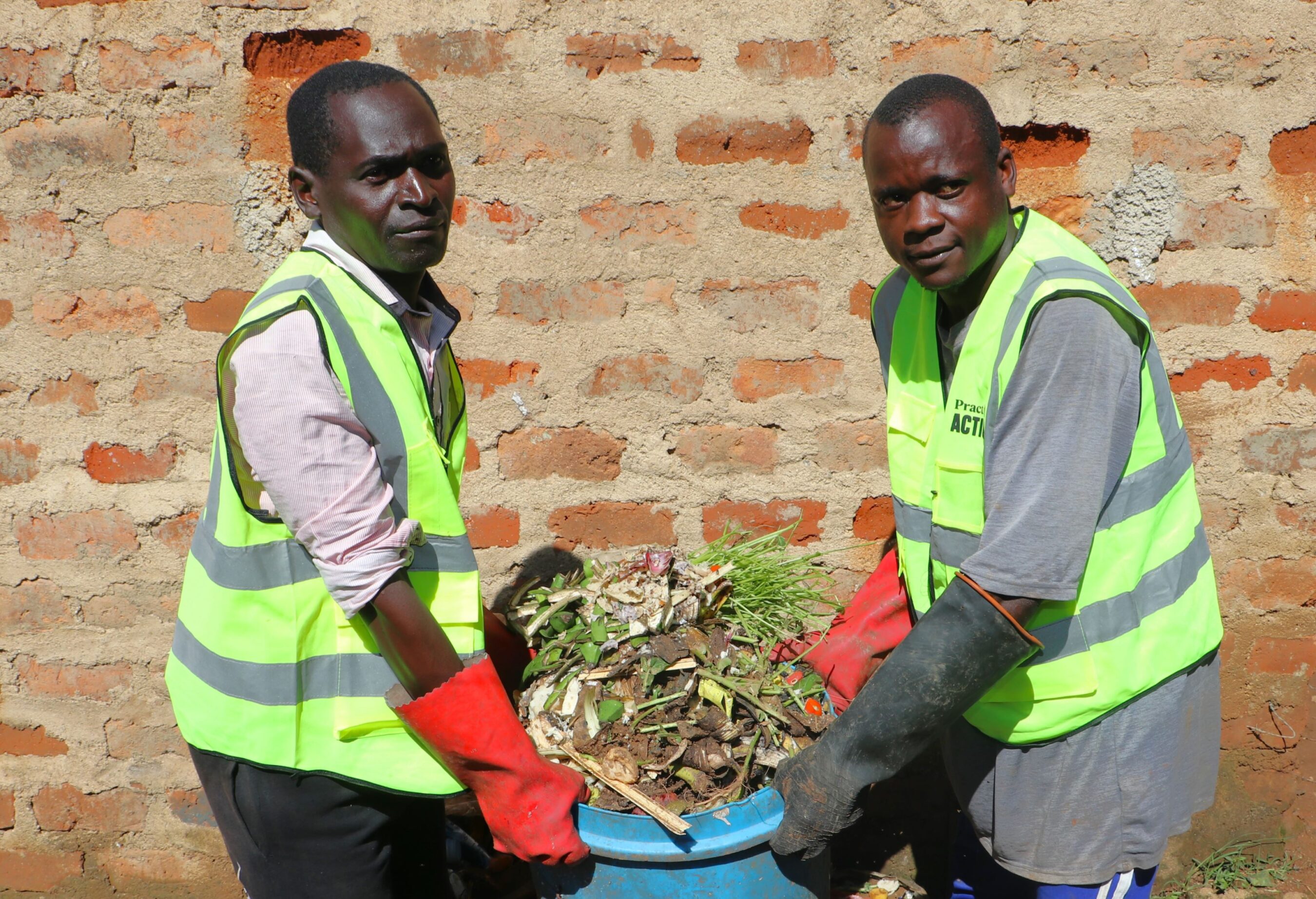 Two men wearing reflective vests and red gloves lift a blue container filled with plant debris in front of a brick wall.