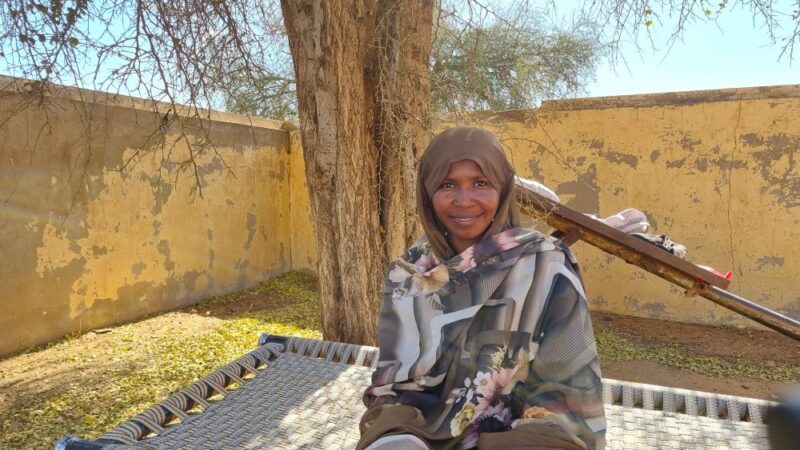 A woman sitting on a bench with a rifle, aiding North Darfur.