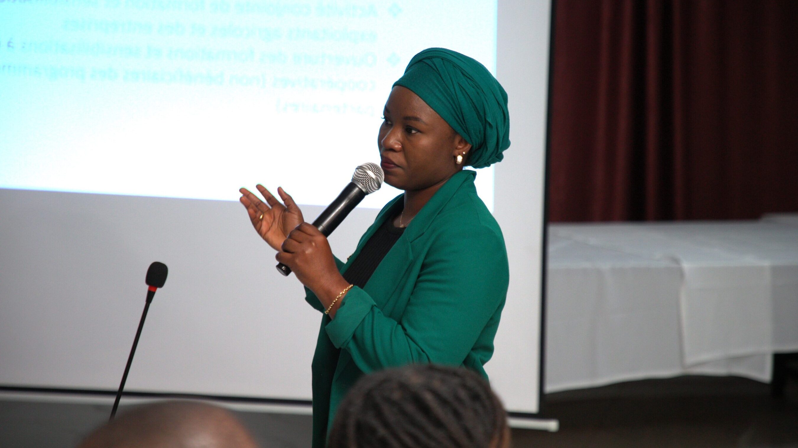 A woman in a green outfit and turban speaking at a conference with a microphone in her hand and a projection screen in the background.