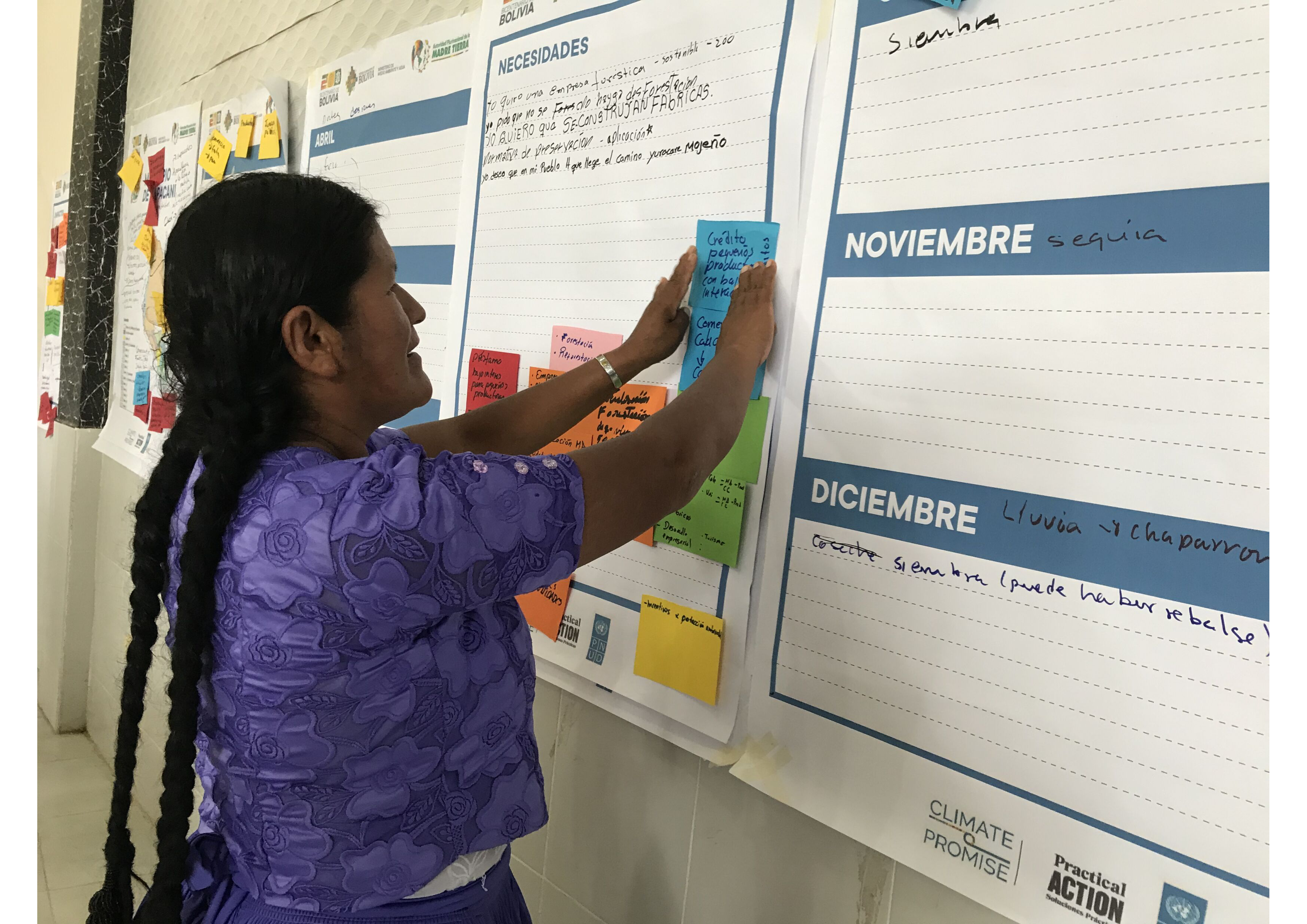 A woman is placing a sticky note on a whiteboard that is being used for event planning or project management, with various sections marked by months such as november and december written in spanish.