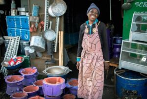 A joyful woman in a brown apron stands in front of a shop with colorful pots and kitchen utensils.