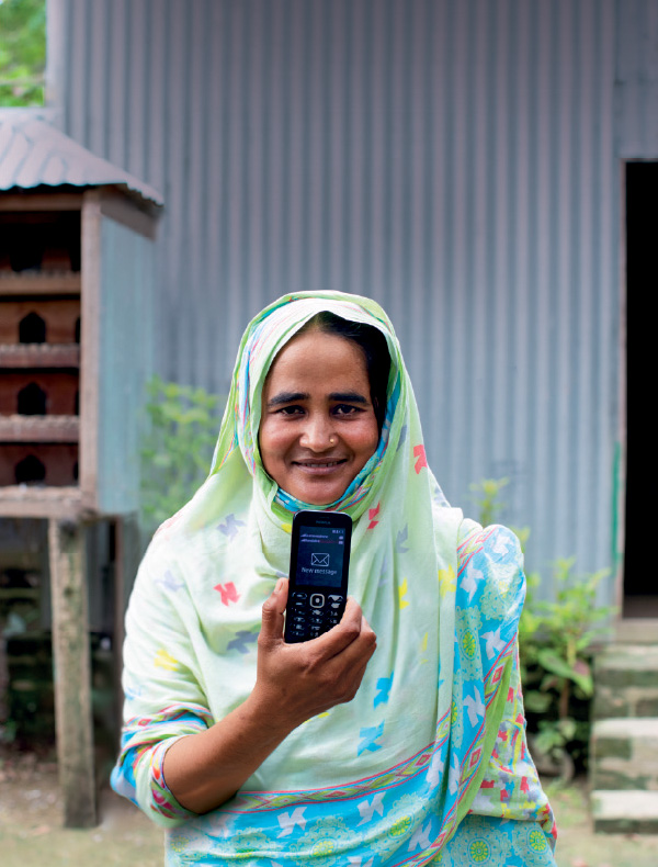 A smiling woman in a floral hijab holds up a mobile phone, standing in front of a house with a wooden beehive.