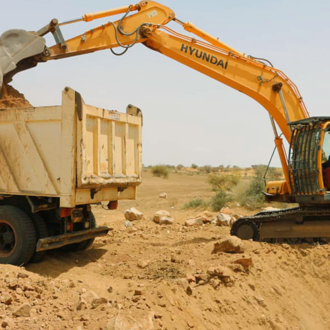 An orange Hyundai excavator loading dirt into the bed of a dump truck at a construction site with a barren landscape in the background.