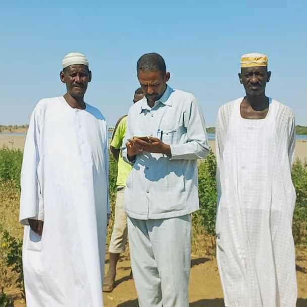 Three men in traditional attire stand outdoors, with one in the center looking at a mobile phone.