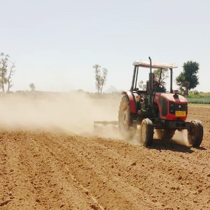 A red tractor plowing a dry field, raising a cloud of dust under a clear sky.