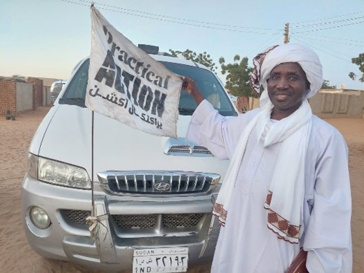 A man in traditional white sudanese attire holding a banner next to a white suv in a dusty outdoor setting.
