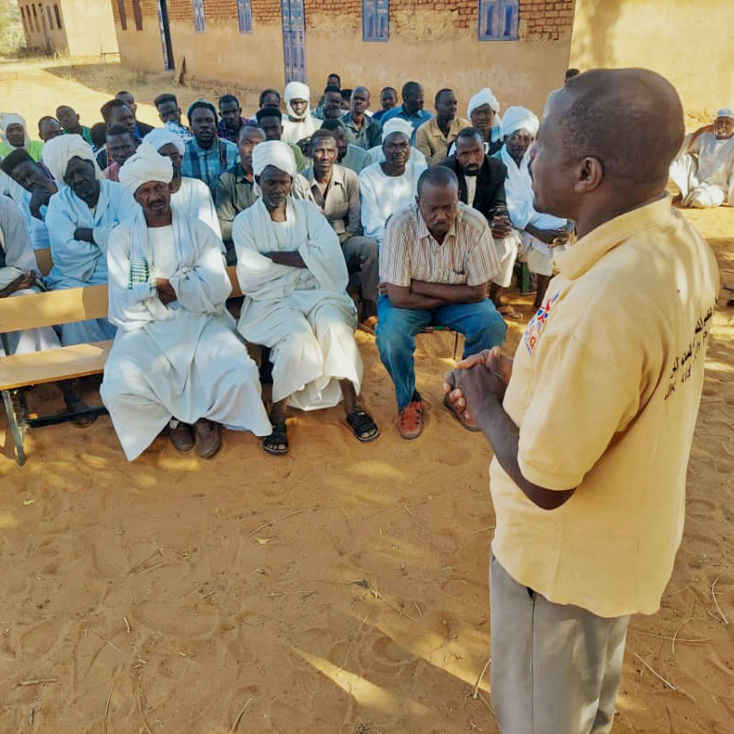 A man speaks to a group of seated men wearing traditional white robes and turbans in a sandy outdoor setting.