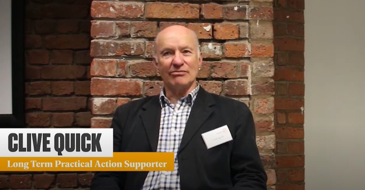 A man named Clive Quick, identified as a long-term Practical Action supporter, sitting in front of a brick wall, wearing a black blazer over a checkered shirt.