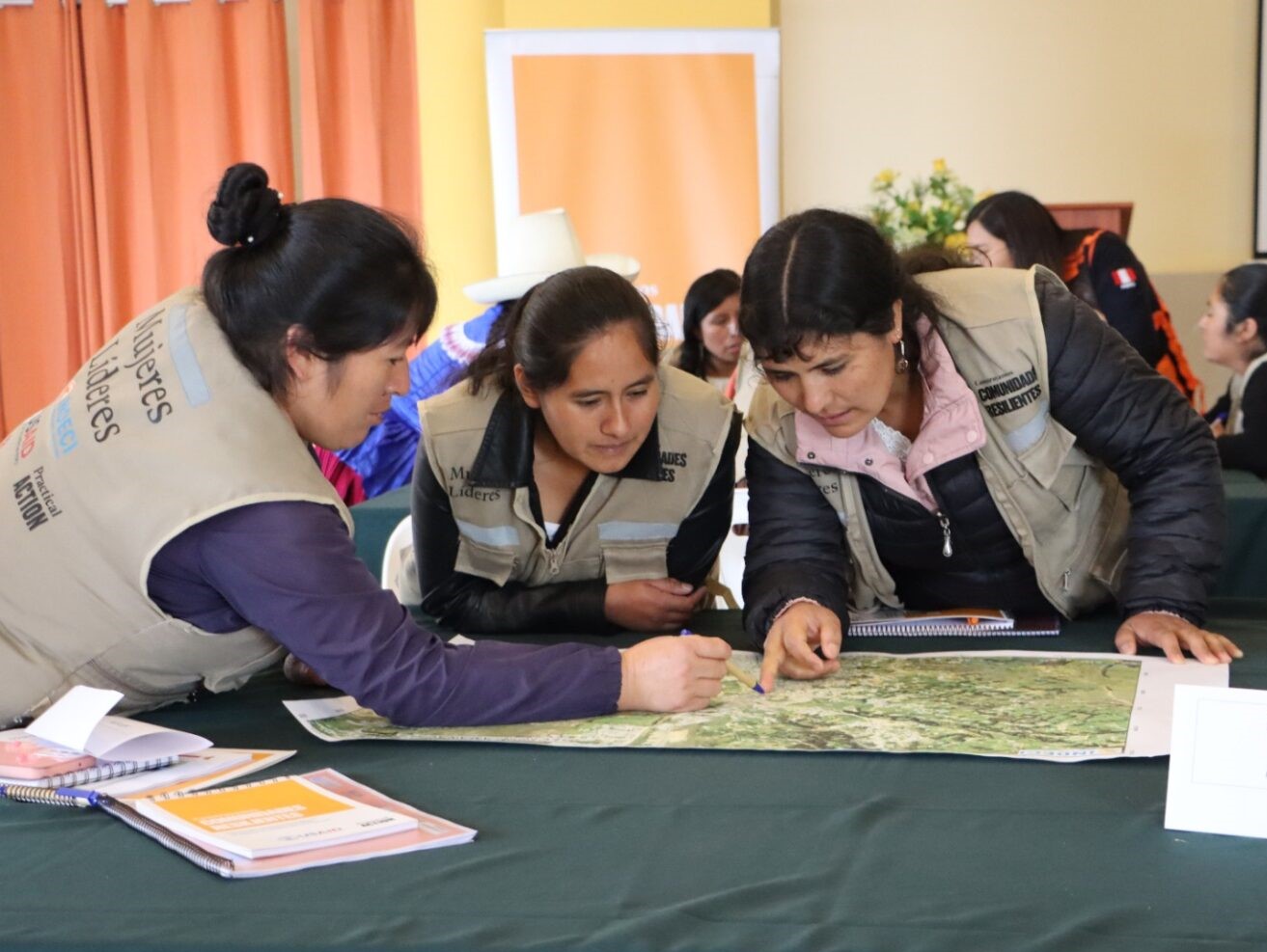 Three individuals in beige vests examine a map on a table, collaborating in a room with orange curtains and other people in the background.