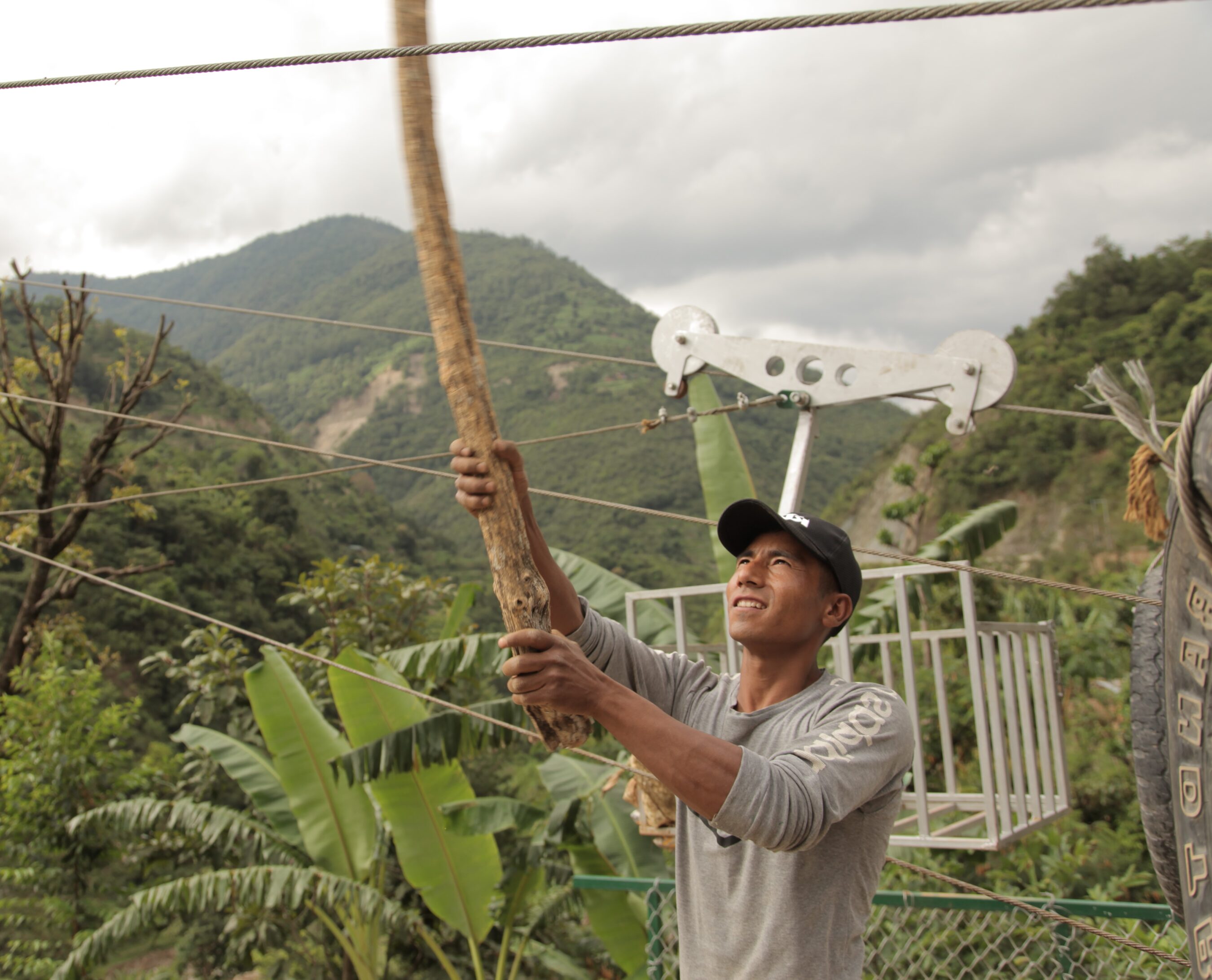 A person in a gray shirt and black cap tightly pulls on a rope in a tropical, mountainous area with a metal structure and lush greenery in the background.