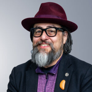 Bill Liao - A man with glasses and a graying beard wears a burgundy hat, black jacket, and purple shirt. He is smiling against a neutral background.