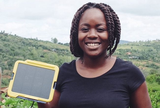 A smiling woman stands outdoors holding a small solar panel device with a hilly landscape in the background.
