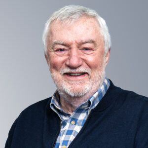 Graham Young - A man with white hair and beard is smiling while wearing a chequered shirt and dark cardigan. The background is gray.