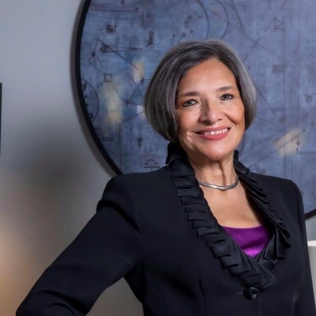 Isabel Studer Noguez - A woman with short gray hair, wearing a black jacket over a purple top, stands in front of a circular abstract artwork, smiling.