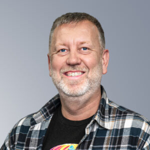 Matt Haikin - A middle-aged man with short hair and a beard smiles at the camera. He is wearing a plaid shirt over a black t-shirt. The background is plain and gray.
