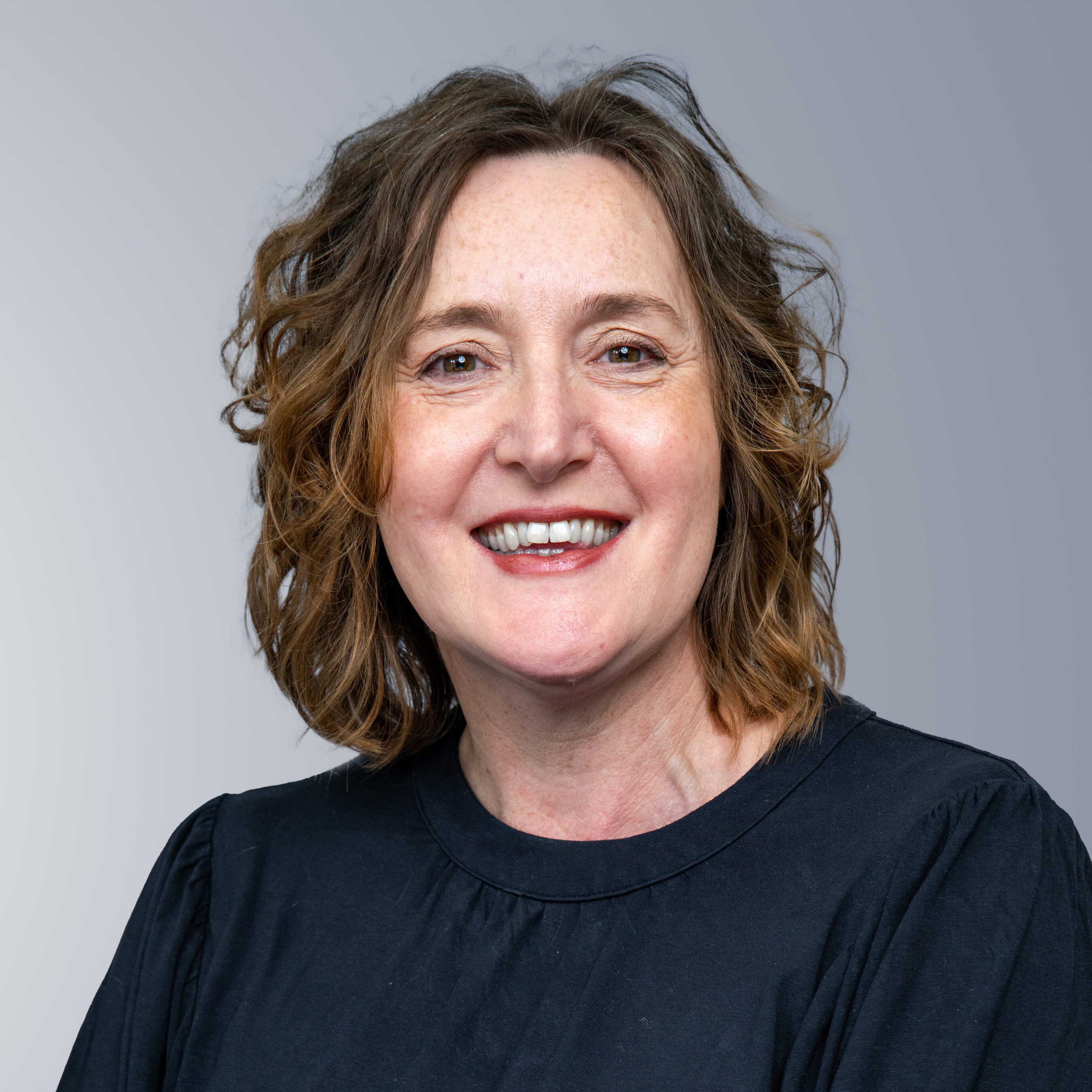Rachel Hudson - A woman with wavy brown hair and a black shirt smiles at the camera against a grey background.