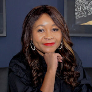 Sazini Mojapelo - A woman with long, dark hair and wearing earrings and a black top is smiling and resting her chin on her hand. She is seated in front of a dark-colored background with artwork.