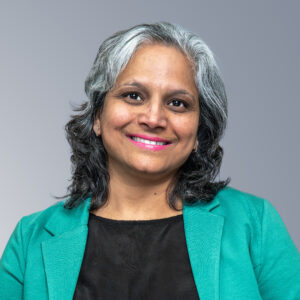 Shivani Wadhwa - A person with gray and black hair smiles, wearing a green jacket over a black top, against a gray background.