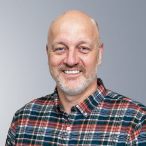 Simon Hotchkin - A bald man with a beard smiling, wearing a red, blue, and gray plaid shirt, against a gray gradient background.