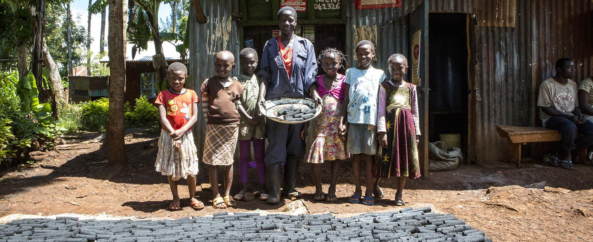 A person stands with a tray of charcoal briquettes, surrounded by six children, in front of a metal building. Charcoal briquettes are laid out on the ground, drying. Trees are visible in the background.