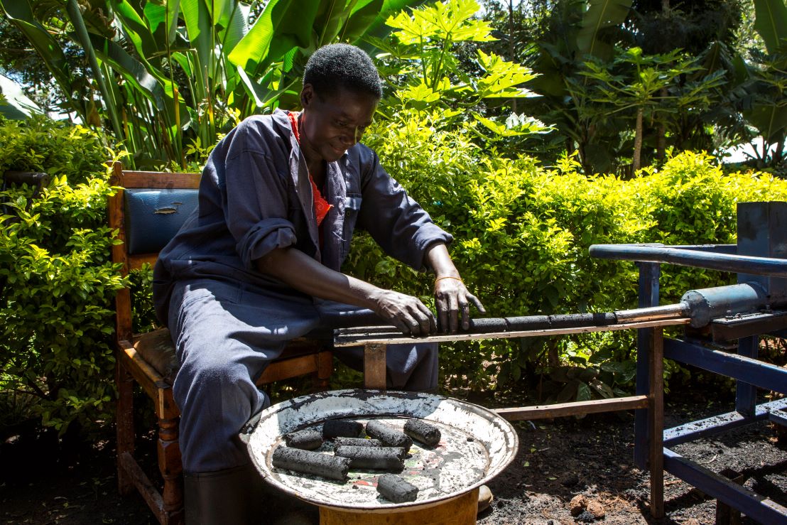 A person in blue overalls makes eco-friendly charcoal briquettes by rolling a mixture in a mold. Lush green foliage surrounds the outdoor workspace.