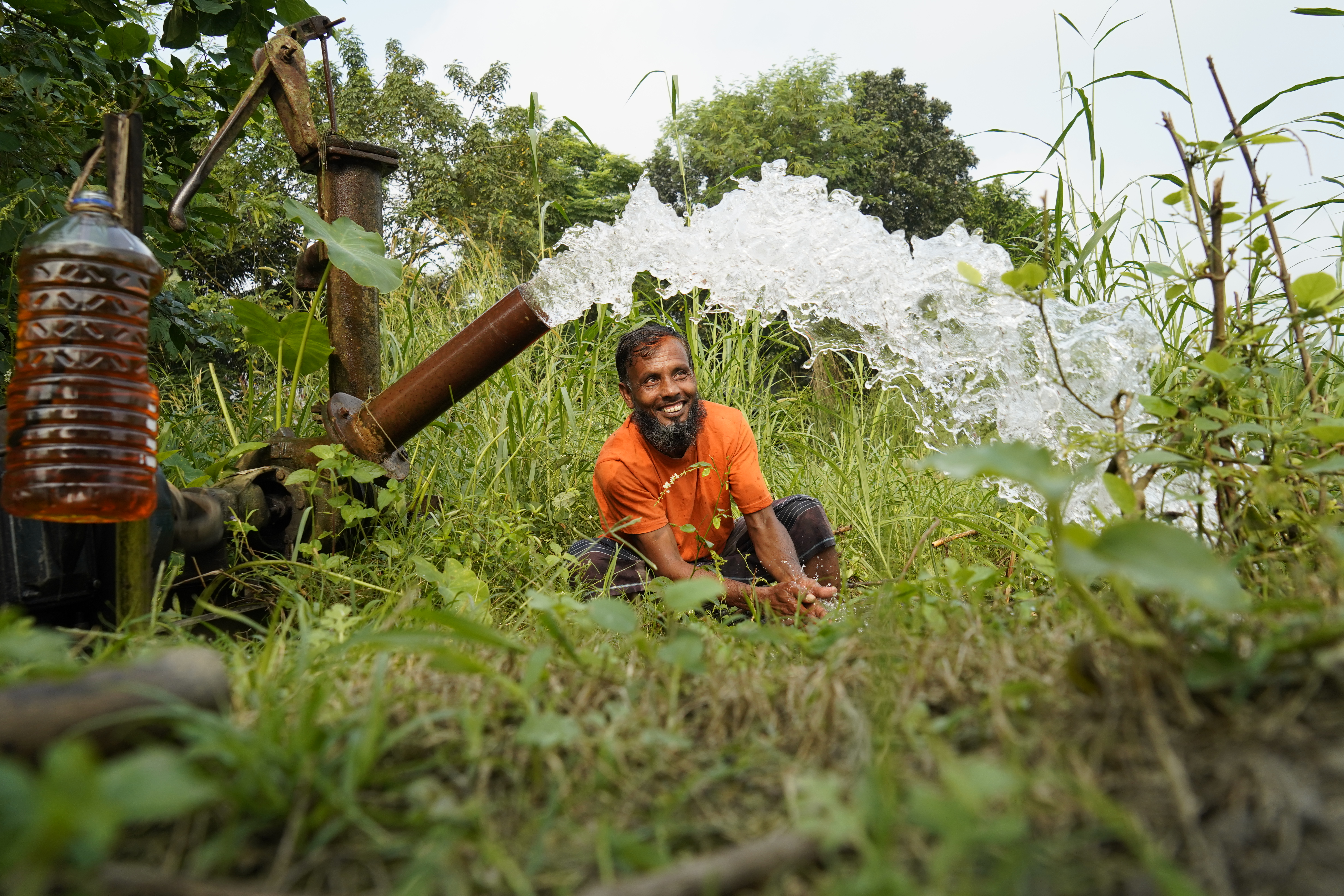 Kolom, a man in an orange shirt kneels on grass by a pyrolysis oil powered water pump, smiling as water gushes out into the air; greenery and trees surround him.
